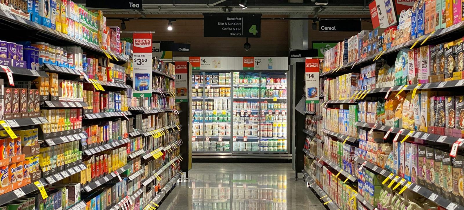 商品の並んだ店内の通路。左右の棚に食品が積まれ、突き当たりに冷蔵ケースが見える。